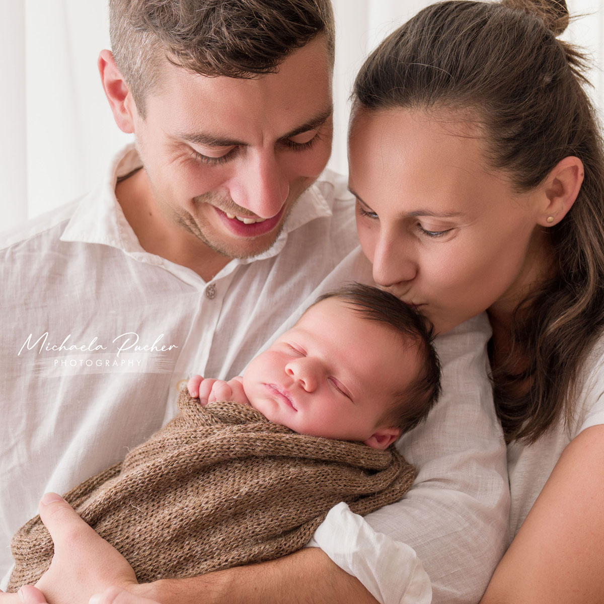 Mama und Papa halten ihr schlafendes Neugeborenes im Arm; die Mama küsst das Baby sanft auf den Kopf, während der Papa lächelnd auf sein Kind blickt.